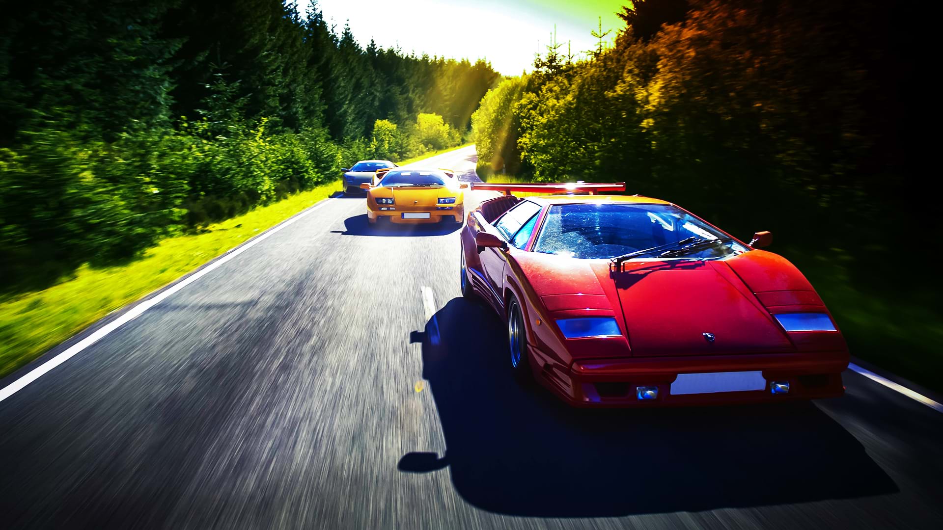Un convoi Lamborghini coloré : Countach 25th Anniversary, Diablo 6.0 et Murciélago, court le long d'une route pittoresque par une journée ensoleillée.